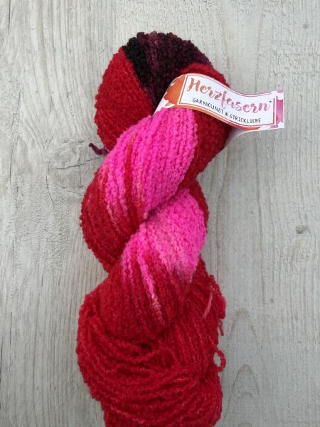 Ein handgefärbter Bouclé-Garnstrang in kräftigem Rot und leuchtendem Pink mit dunklen Akzenten liegt auf einem hellen Holzuntergrund. Die Oberfläche zeigt die typische schlaufenartige Struktur des Garns.